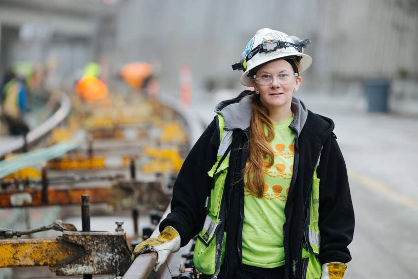 Ali Crespo, graduate of the TRAC pre-apprenticeship program, wearing a green construction vest, hardhat with headlamp, safety glasses and work gloves, stands with her hand on raised rail tracks being installed.
