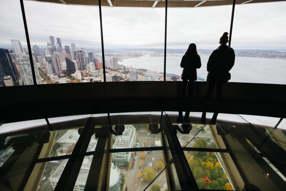 Two people stand on the new glass floor of the Space Needle, looking out the windows toward downtown Seattle.