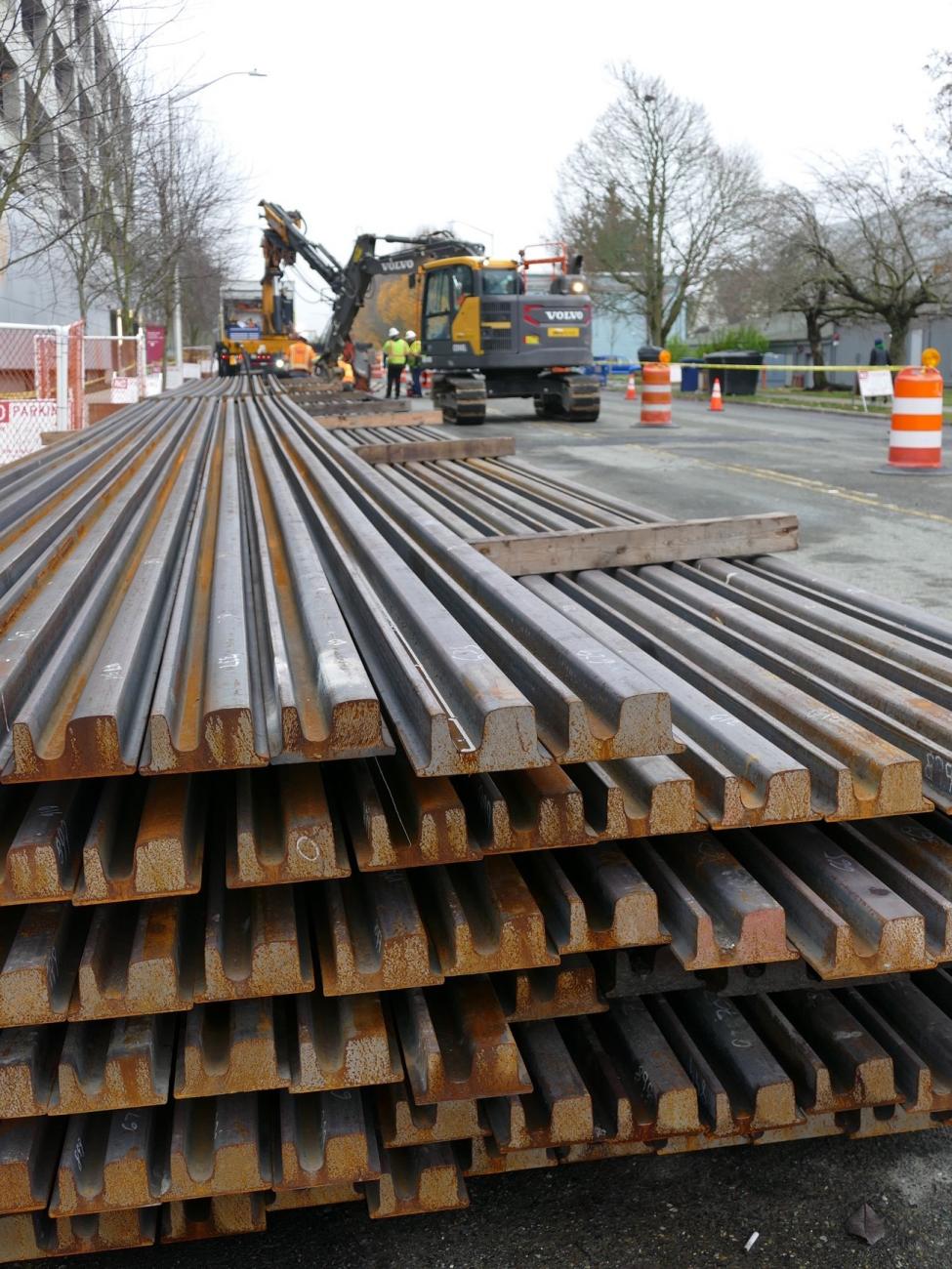 Stacks of rail are pictured, with construction equipment in the background.