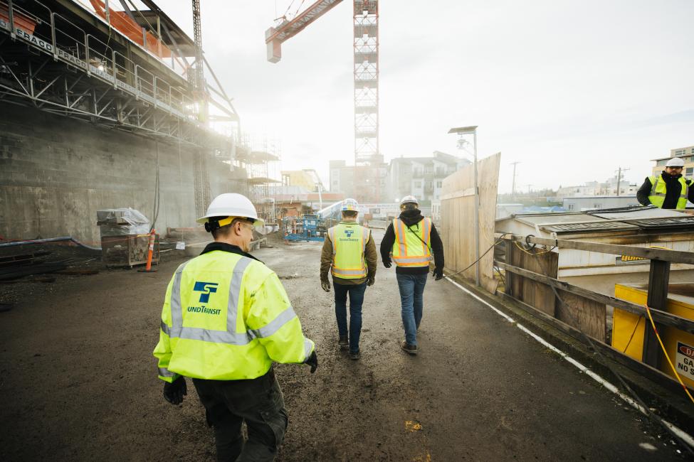 Three men in yellow vests walk toward the construction site at Roosevelt Station.