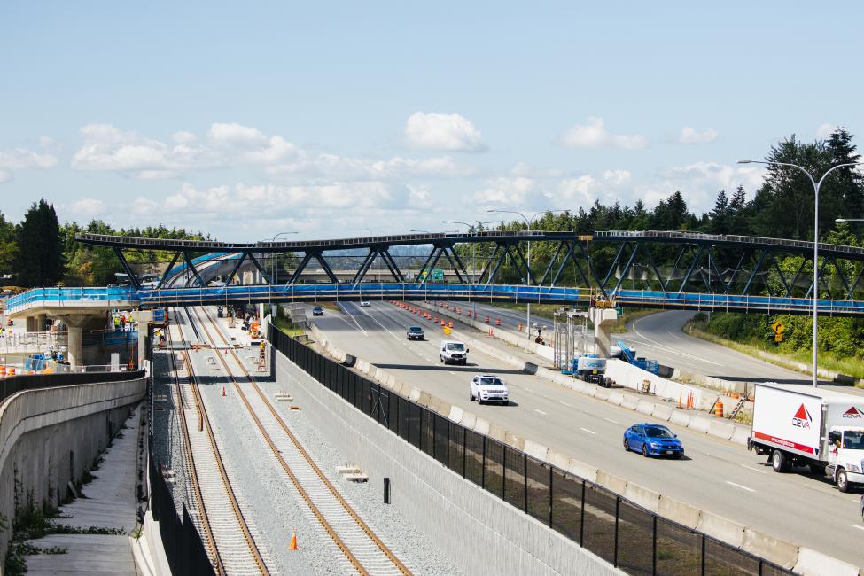 A large bridge crosses over highway SR 520 in Redmond. Several cars are driving on the road. New tracks for light rail can also be seen on the south side of the freeway.