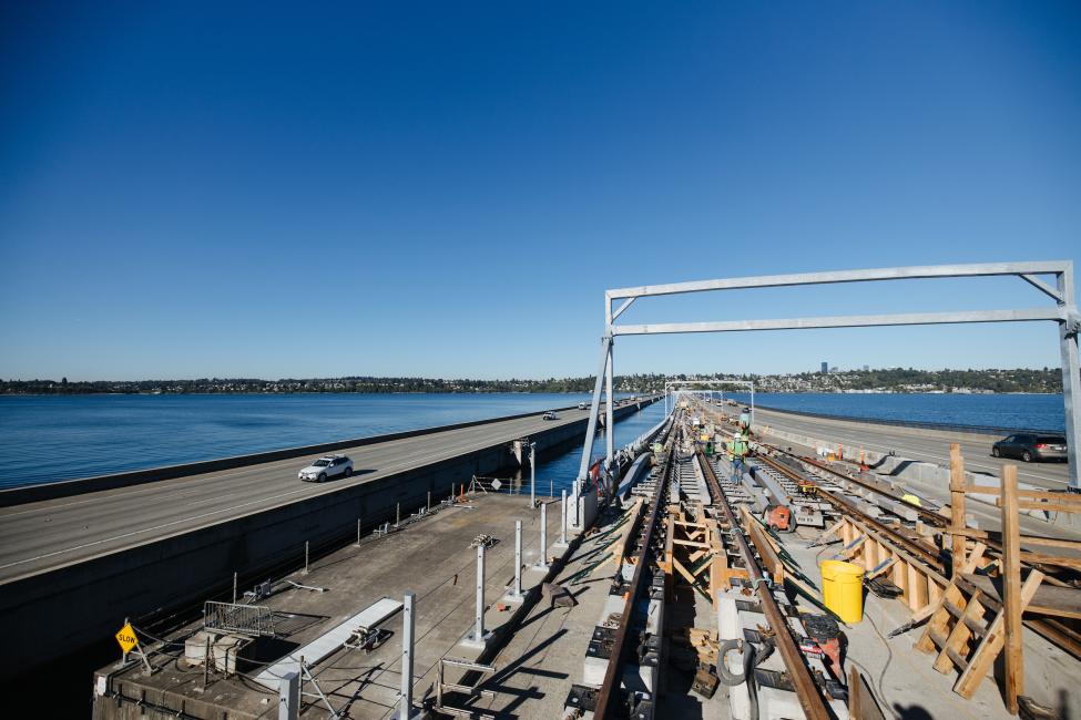 Light rail tracks are installed in the center lanes of the I-90 floating bridge, with Lake Washington visible on the left side of the photo.