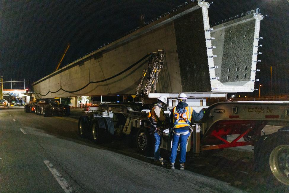 Workers stand next to a very large concrete girder span, sitting on a truck bed before getting ready to be lifted up.
