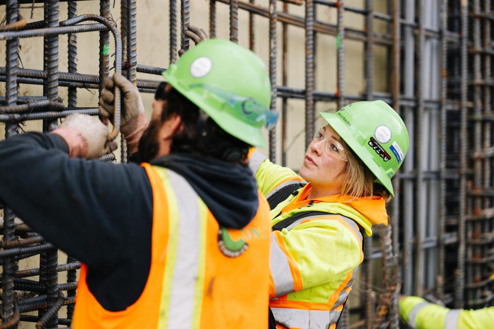 Two construction workers in hard hats and vests work on a rebar cage.