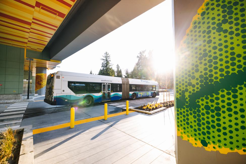A Sound Transit Express bus passes underneath the elevated South Bellevue Link light rail station platform as rare fall sunshine pours directly into the camera lens.