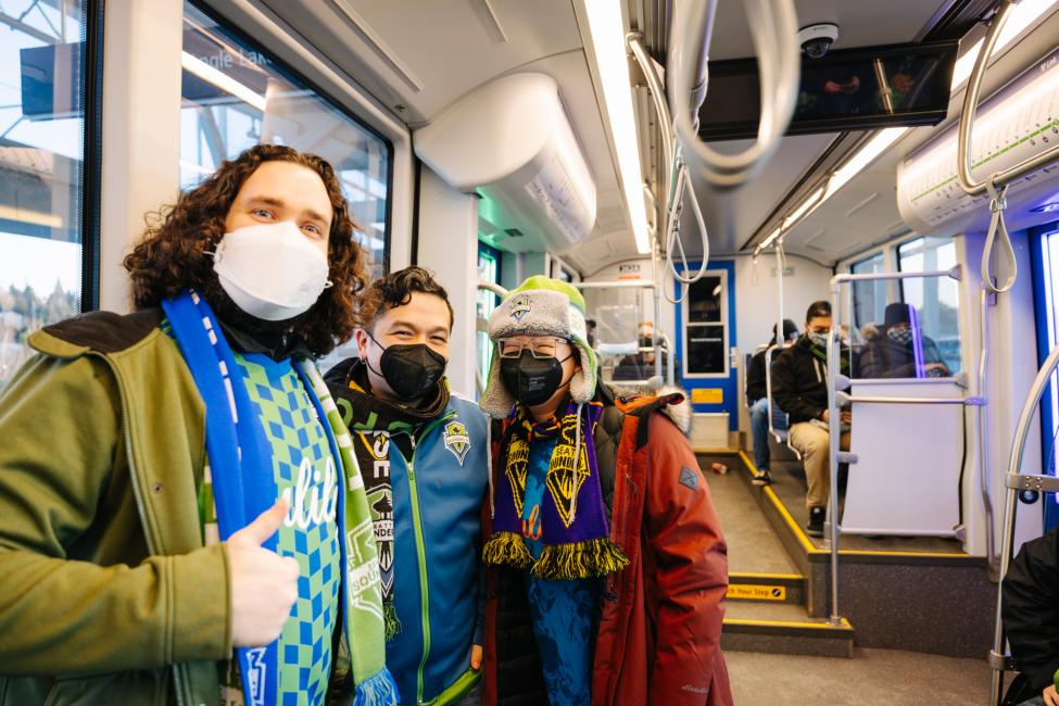 Three Sounders fans in jerseys, scarves and face masks ride a Link train.