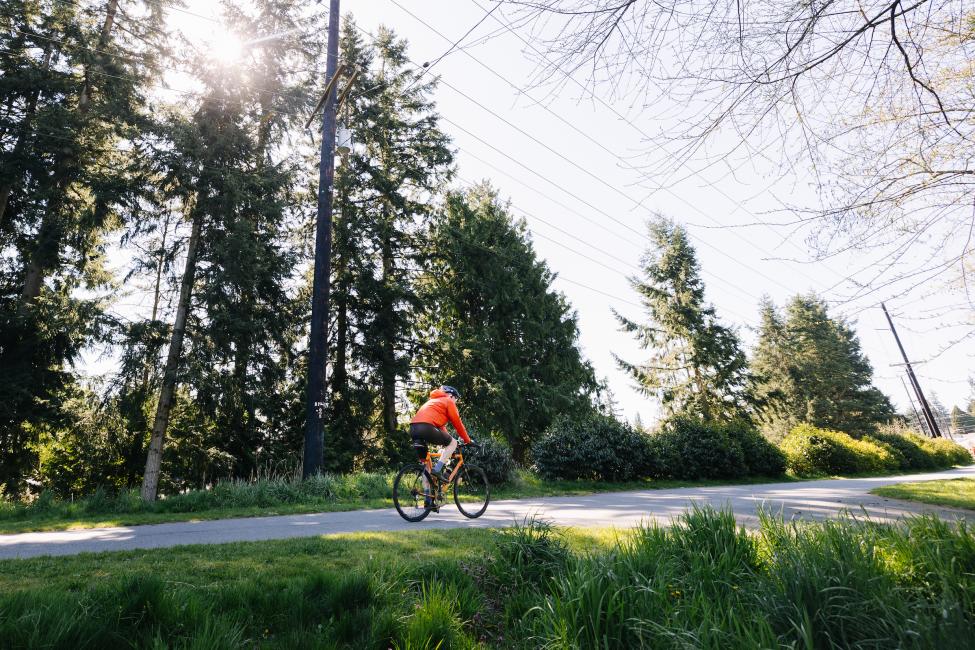 A cyclist rides on the Interurban Trail on a sunny day, with green trees in the background.