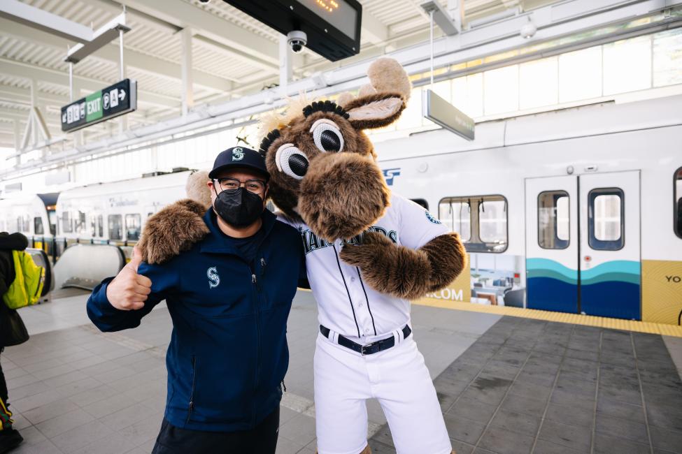 The Mariner Moose hugs a passenger wearing a baseball hat and face mask at Northgate Station.