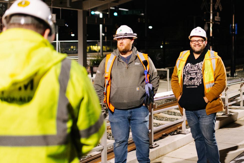 Three people wearing orange vests and hard hats stand on a train platform at night.