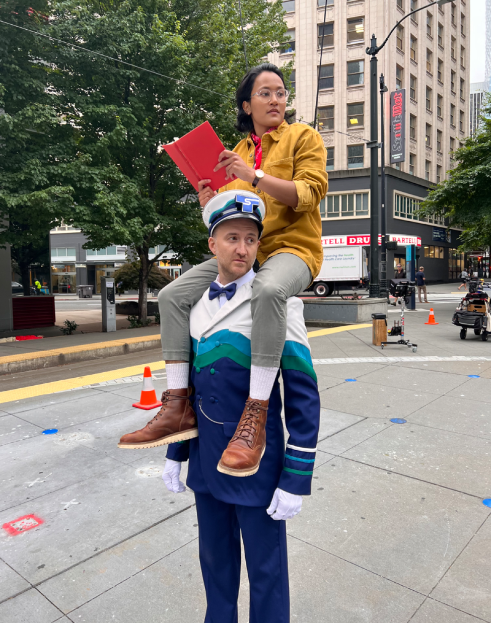 An actor in an orange jacket sits on the shoulders of the Sound Transit conductor character, who is wearing blue, green, white and a hat.