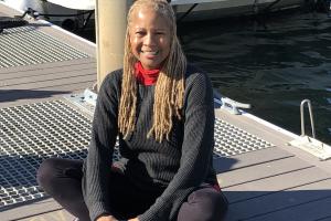 Galen sits cross-legged on a boat dock, wearing a gray sweater over a red turtleneck.