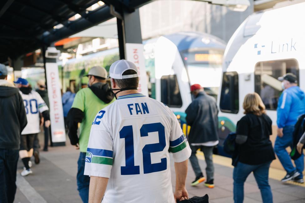 A Seahawks fan in a '12' jersey walks on a Link platform, with a train in the background