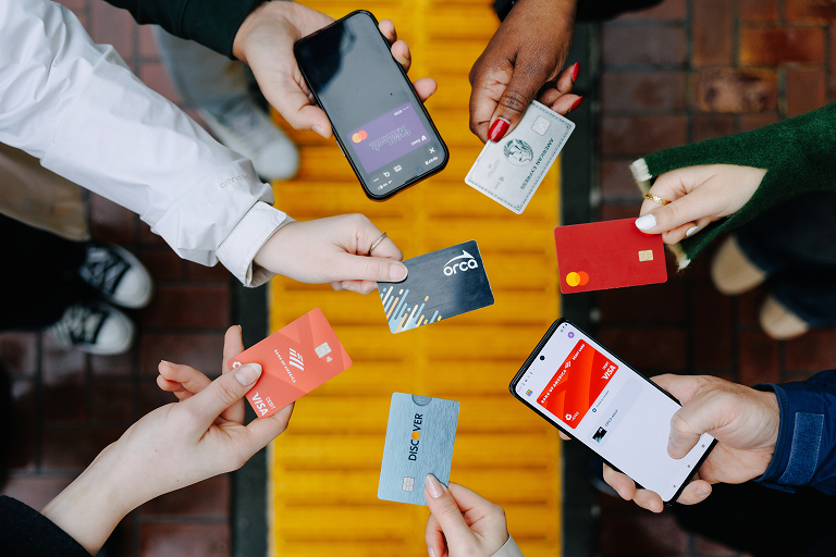 Photo of hands holding various forms of payment cards and phones with an Orca card in the middle