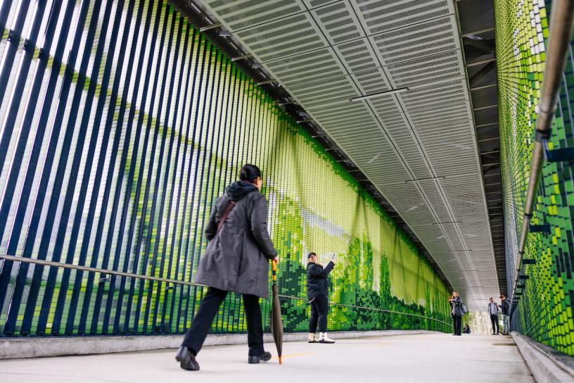 People walk across a pedestrian bridge, which is encased in green panels