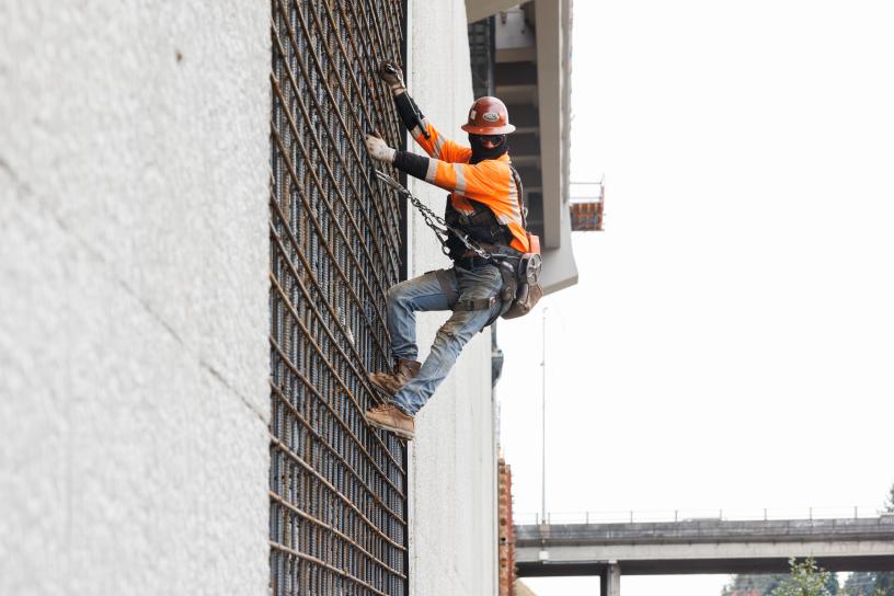 A worker in an orange jacket and a harness climbs a wall at a construction site
