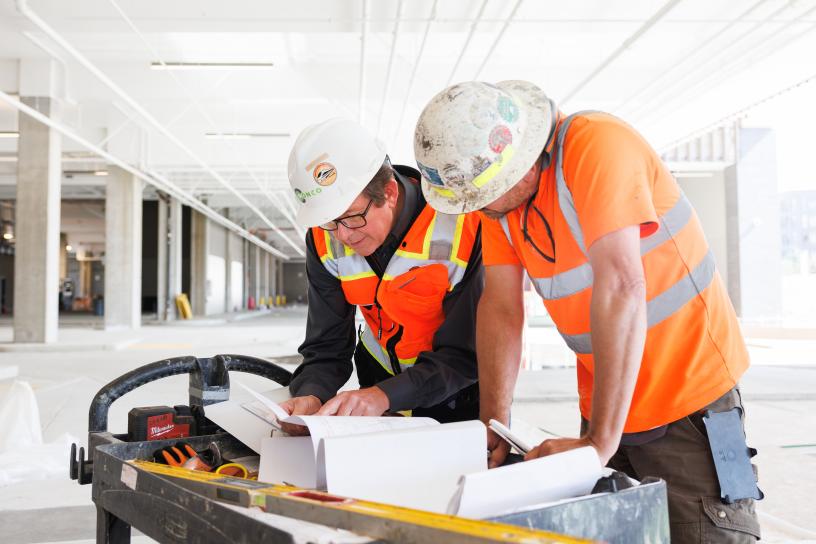 Two construction workers in orange shirts and hard hats look at documents on a table