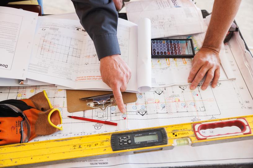 Fingers point at construction plans. Also visible are a ruler, calculator, safety googles and gloves and a red pen