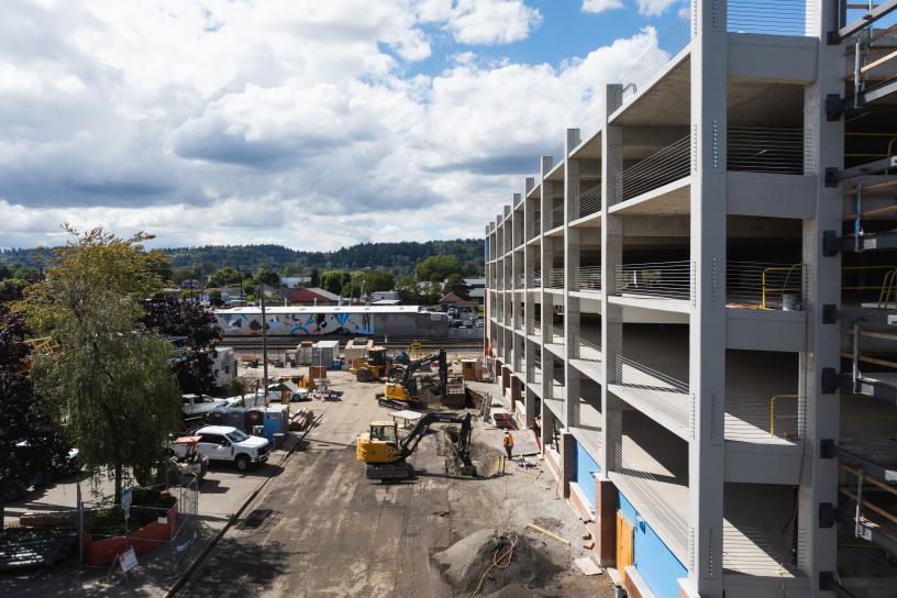 A multi-level parking garage under construction at Auburn Station. The concrete structure includes a prominent blue elevator tower, exterior stairways, construction equipment, fencing, and active work zones surrounding the site.