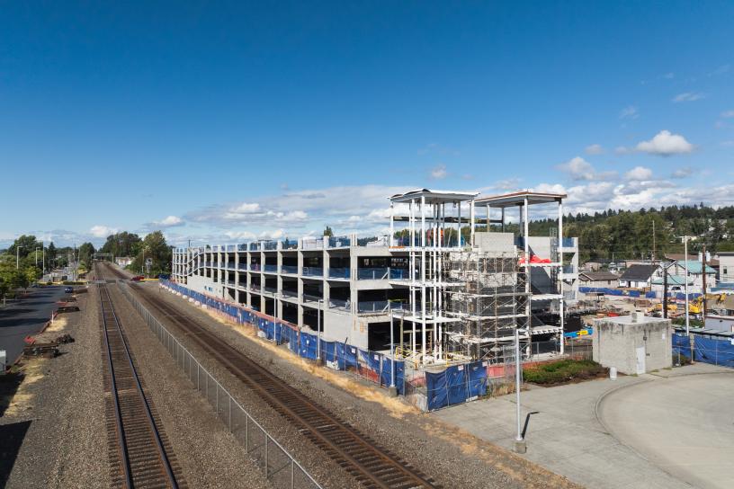 Aerial view of the Kent Station parking garage under construction adjacent to the Sounder platform. The multi-level concrete structure is largely complete, with crews performing site work, roadway improvements, and finishing activities around the station area.