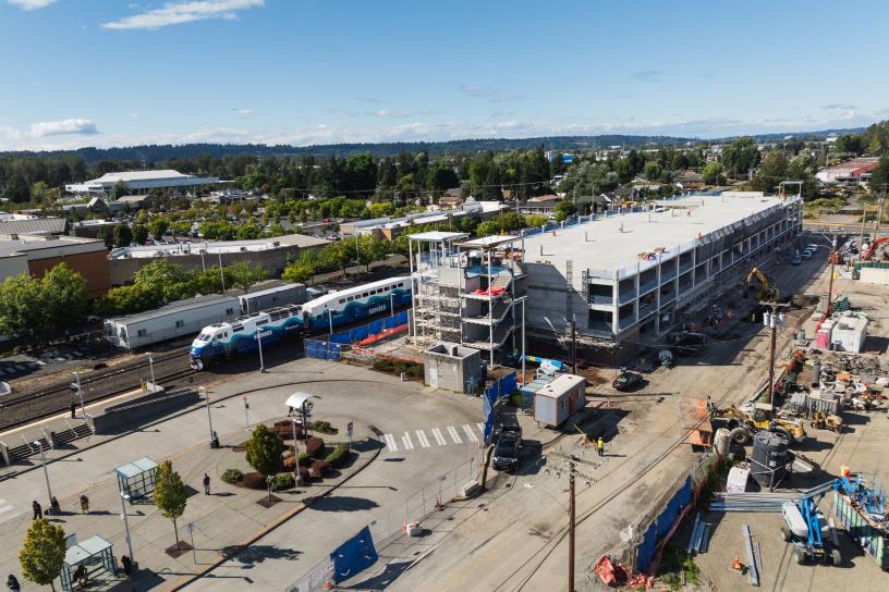 The Kent Station parking garage under construction alongside railroad tracks. The multi-level concrete structure includes a developing elevator tower, scaffolding, construction fencing, and active work areas adjacent to the station platform.