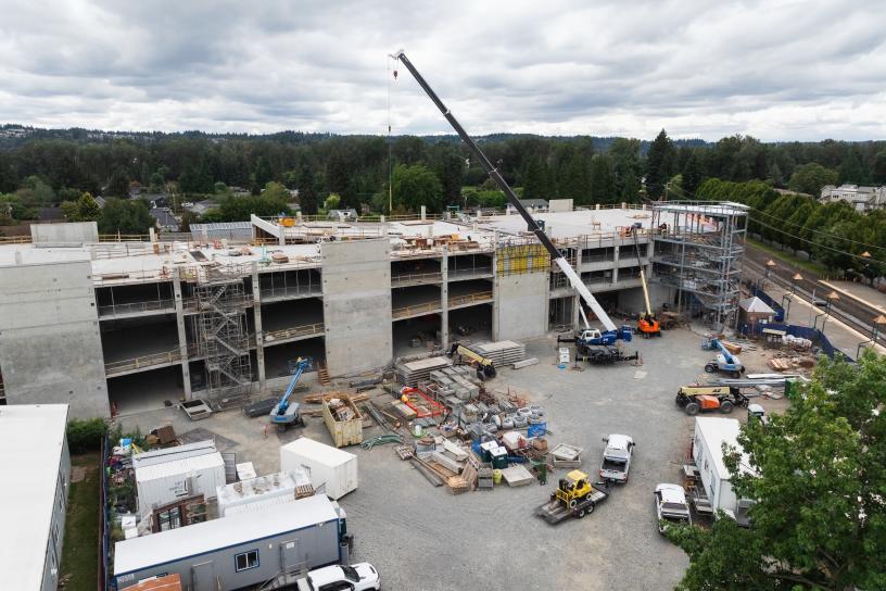 An aerial view of the Sumner Station parking garage under construction. The multi-level concrete structure is taking shape next to the Sounder station platform, with construction equipment, materials, and a freight train visible alongside the rail corridor.