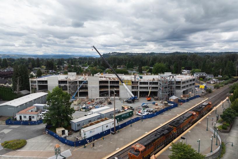 Aerial view of the Sumner Station parking garage construction site showing the nearly completed concrete structure, construction vehicles, materials staging areas, and workers building the upper levels adjacent to the rail station.