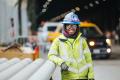 Trai Daniel, graduate of the ANEW pre-apprenticeship program, wears a yellow safety jacket, blue hard hat with a headlamp and safety glasses, leans against a stack of pipe while smiling for the camera.