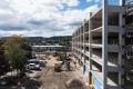 A multi-level parking garage under construction at Auburn Station. The concrete structure includes a prominent blue elevator tower, exterior stairways, construction equipment, fencing, and active work zones surrounding the site.