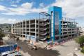 View of the Auburn Station parking garage under construction showing finished portions of the concrete structure alongside active roadway and utility work. Construction vehicles, materials, and work areas are visible adjacent to the building.