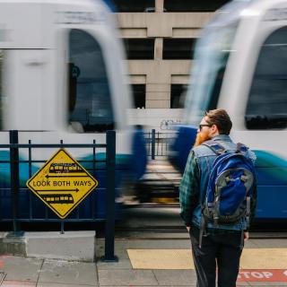 A passenger waits for a Link train to pass before crossing. A yellow sign to his left reads 'look both ways.'