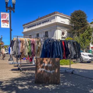 Sandwich board sign for Shop Conmigo out on the sidewalk in the sunshine in front of a rack of clothes