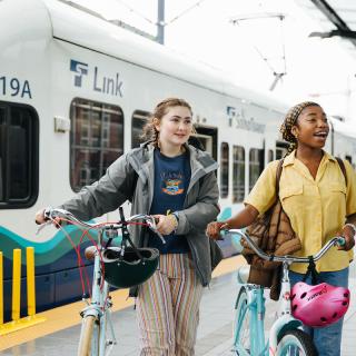 Two teenage girls with bikes walk on the platform at Othello Station.