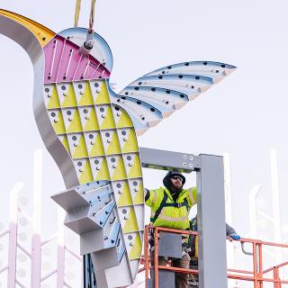 A construction worker on a hydraulic lift puts the finishing touches on a new art installation of a colorful neon-lit hummingbird at Lynnwood Transit Center.