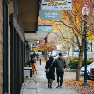 Two people walk on a sidewalk in downtown Burien amidst trees with orange and yellow leaves
