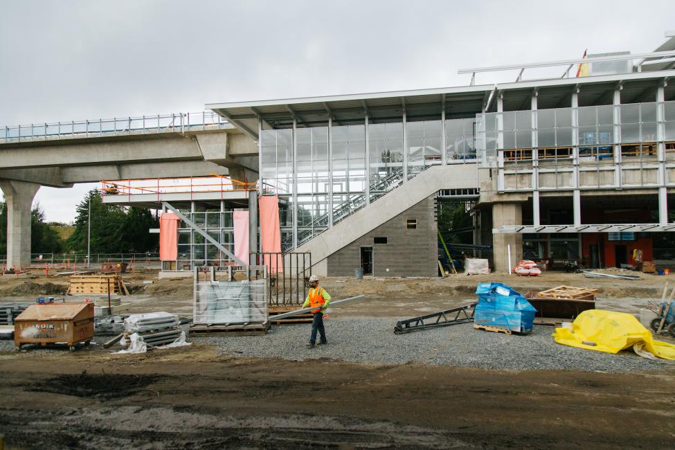 A view of Northgate Station, looking west. A construction worker carries a steel beam in the foreground.