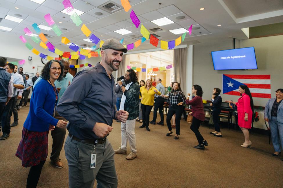 Sound Transit employees, including Jefferson Rose, dance at a Latinx Heritage Month kickoff celebration in a large room decorated with colored tissue paper banners and flowers.