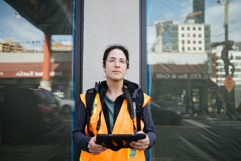 A woman in an orange vest holds a tablet while looking at buildings in Seattle's International District/Chinatown neighborhood.