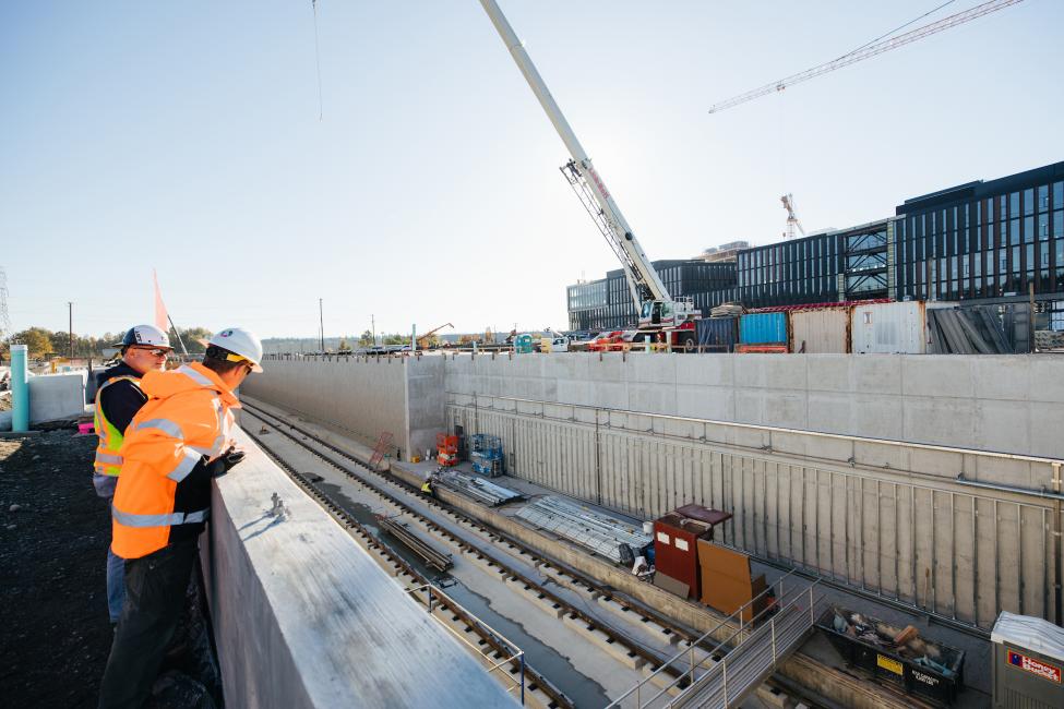 Two men in hard hats peer over a wall to look down into the Spring District's light rail station, currently under construction.