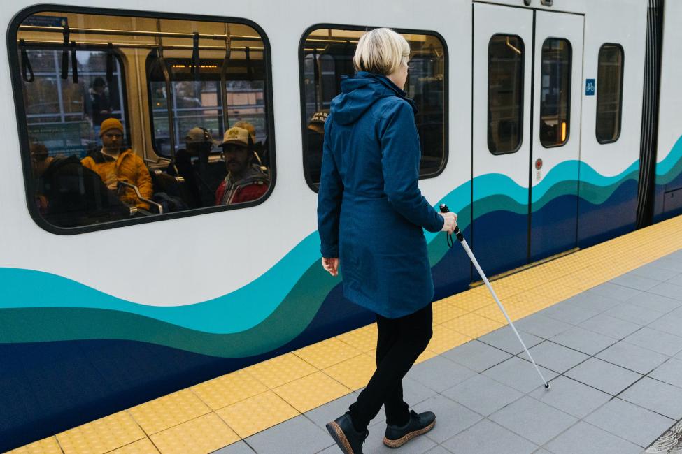 A blind woman uses a cane and tactile warning mat to navigate through a Link station.