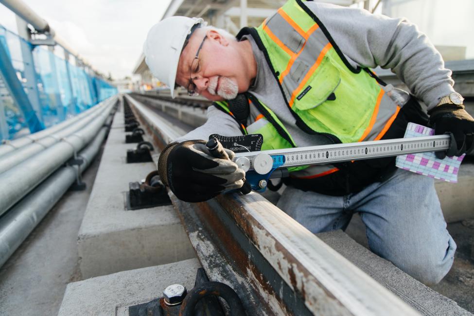 A man in a hard hat and construction vest uses a tool to measure a section of rail at Northgate Station.