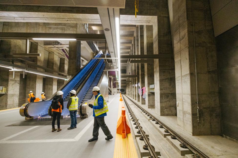 People in hard hats stand on the platform of Roosevelt Station at the base of newly installed escalators.