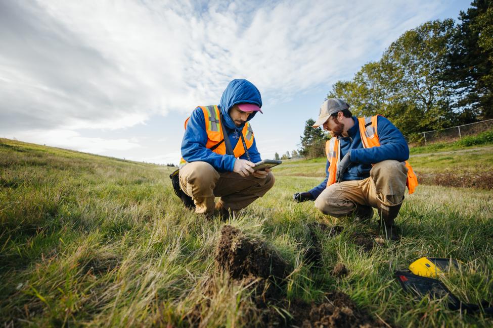 Two men in orange vests kneel in a grass field, looking at fieldwork equipment.