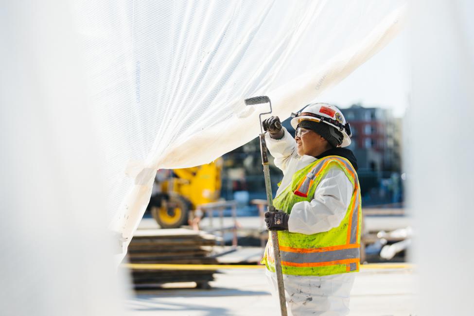 Underneath a large white tarp, a woman uses a big brush to paint at Spring District Station.