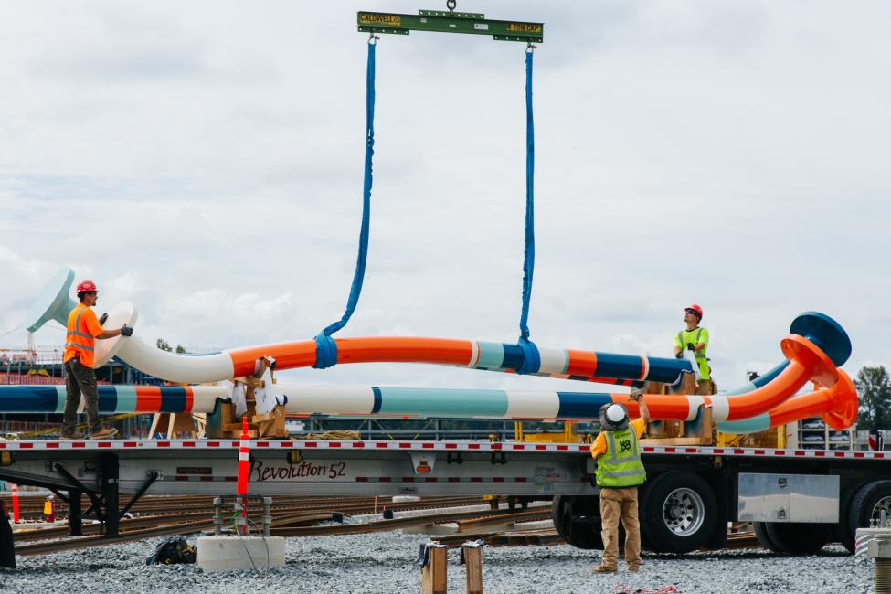 Workers use equipment to lift large sculptures that look like colorful bent nails.