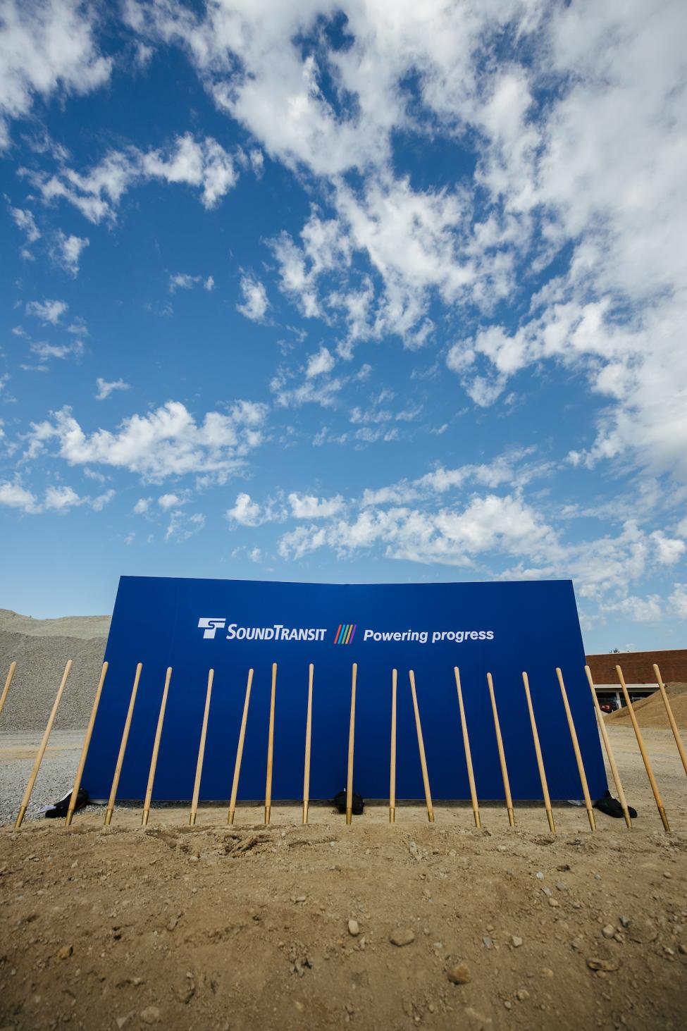 A row of shovels lined up in the dirt, with a blue backdrop that has Sound Transit's logo and the words "Powering progress."