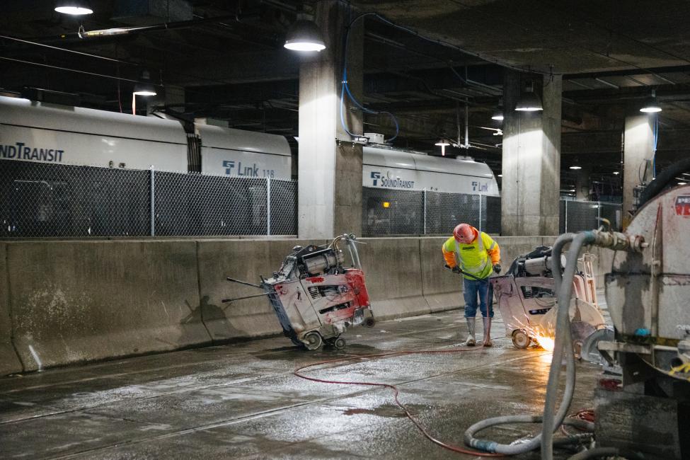 A worker cuts a piece of rail with a big saw at the construction site at International District/Chinatown Station. The top of a Link train can be seen in the background.