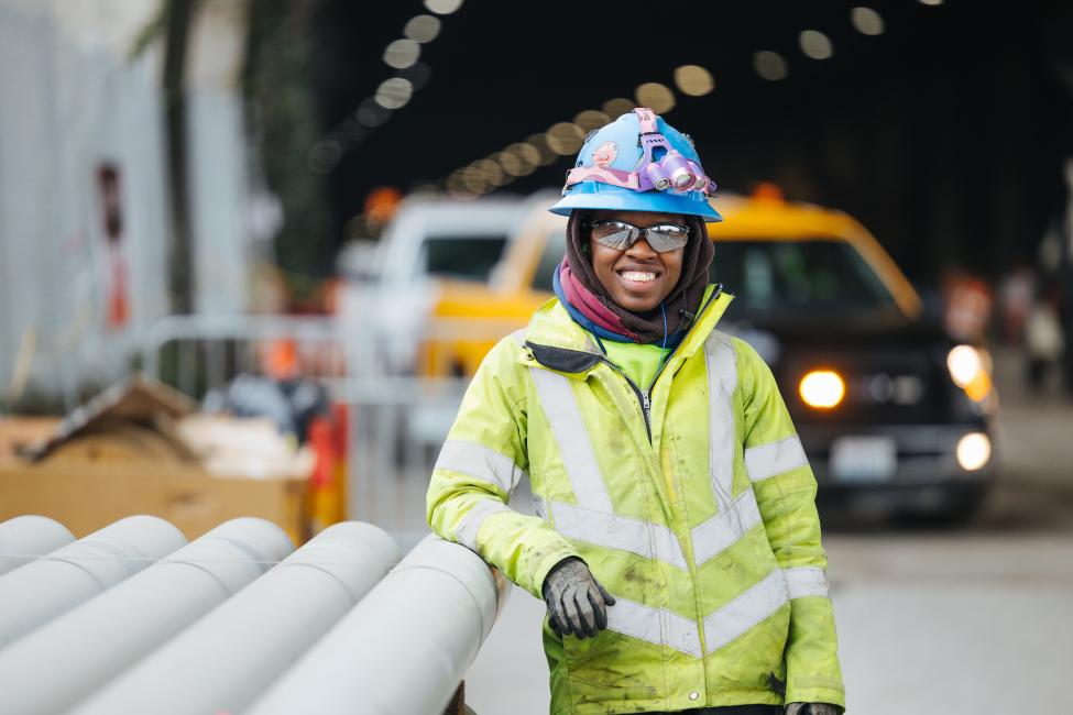 A woman in a yellow jacket, safety goggles and blue hard hat smiles, with construction trucks and equipment in the background.