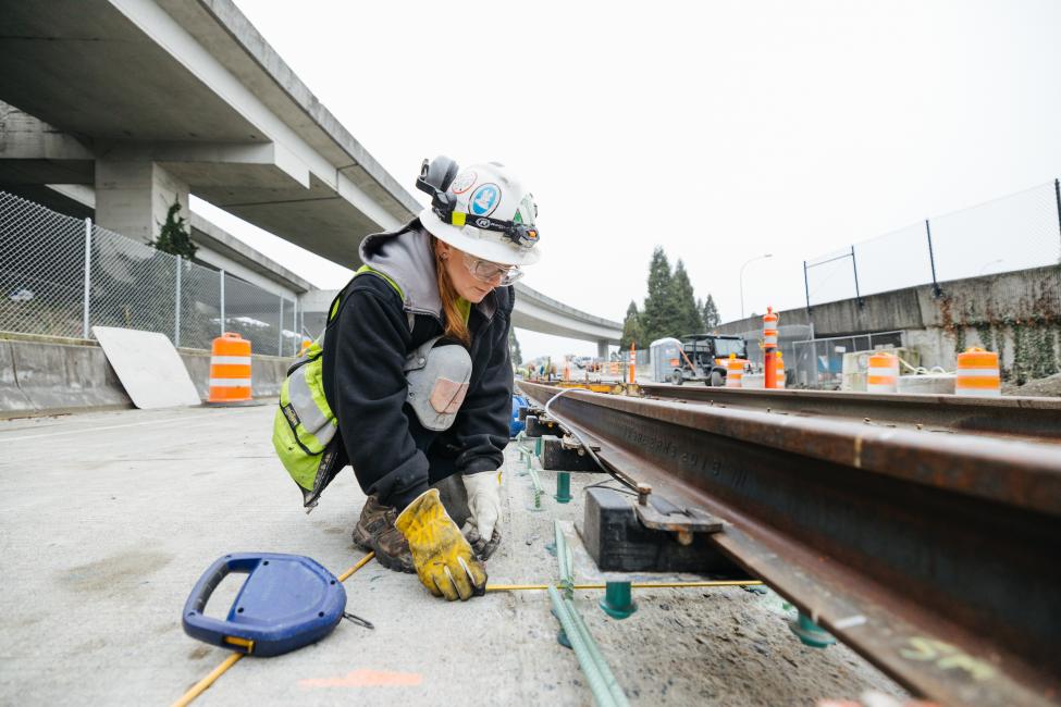 A woman with gloves, a yellow vest and hard hat kneels down to measure a length of cement underneath a rail.