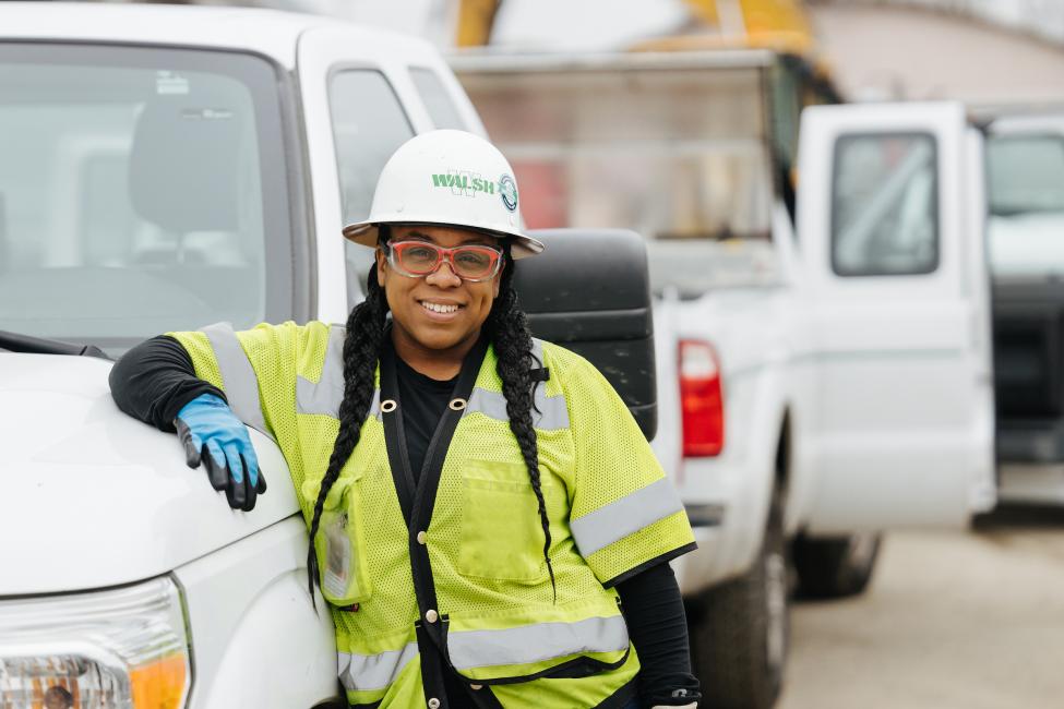 A woman in a yellow vest, hard hat and red glasses under her safety goggles smiles at the camera, with her arm resting on the hood of a pickup truck.