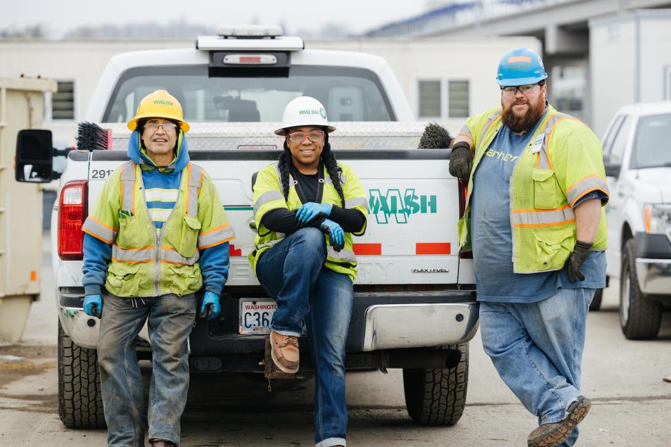Two men and one woman in vests, gloves, hard hats and work boots sit on the back bumper of a pickup truck.